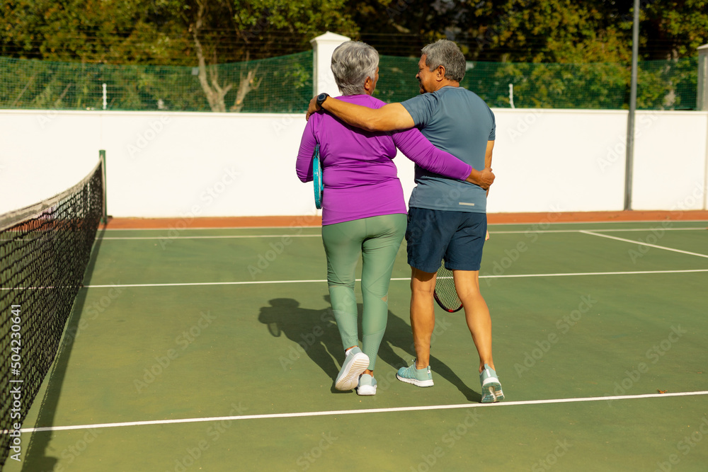 Rear view of biracial senior husband and wife with arms around walking at tennis court on sunny day