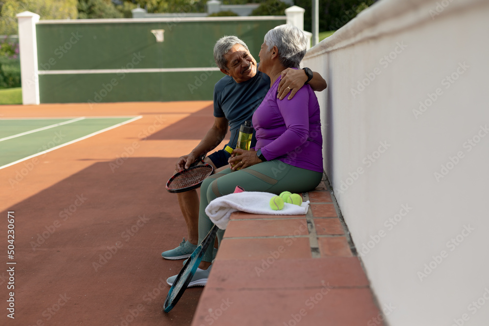 Smiling biracial senior husband looking at wife while sitting on bench at tennis court, copy space Smiling biracial senior husband looking at wife while sitting on bench at tennis court, copy space