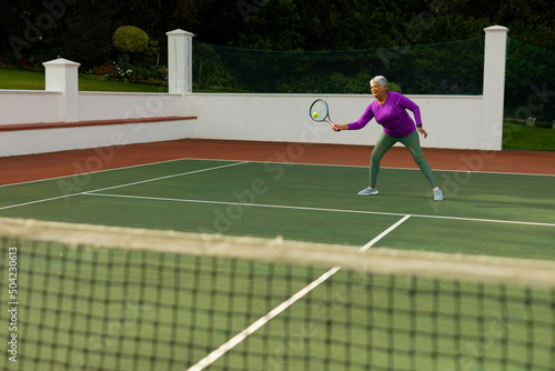 Full length of biracial senior woman hitting tennis ball with racket while playing tennis at court