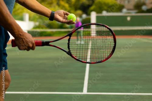 Midsection of biracial senior man holding racket serving tennis ball while playing at tennis court