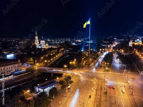 Fotografia Illuminated flag of Ukraine and Holy Annunciation Cathedral on river embankment with transport on bridge across river Lopan at night