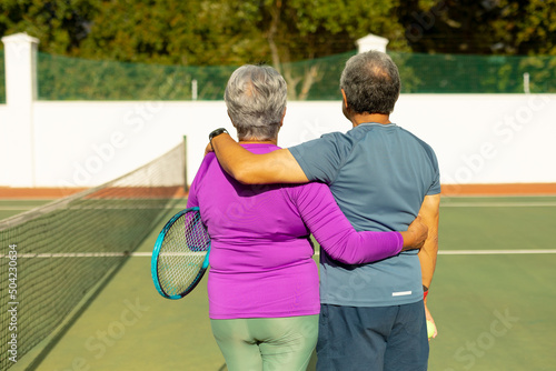 Rear view of biracial senior wife and husband with arms around standing at tennis court on sunny day