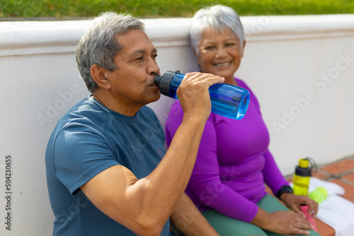 Tired biracial senior man drinking water while sitting with smiling wife on bench at tennis court
