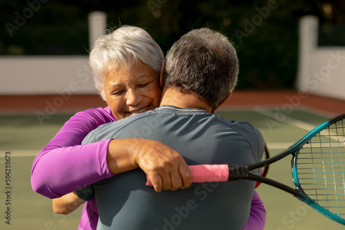 Rear view of romantic biracial senior man embracing smiling wife holding racket at tennis court