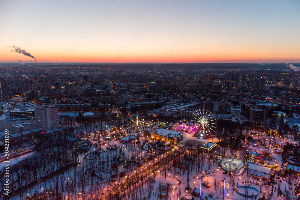 Aerial view on evening city lights, streets, Ferris wheel, entertainments in winter, covered in white snow. Kharkiv city center in sunset colors, amusement Gorky Central Park