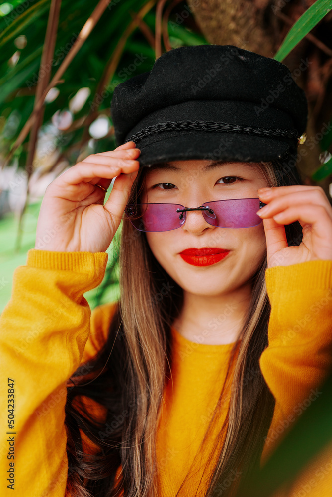 vertical portrait of young asian girl, wearing yellow sweater, purple sunglasses and beret, smiling under palm tree. fashion and beauty vertical portrait of young asian girl, wearing yellow sweater, purple sunglasses and beret, smiling under palm tree. fashion and beauty