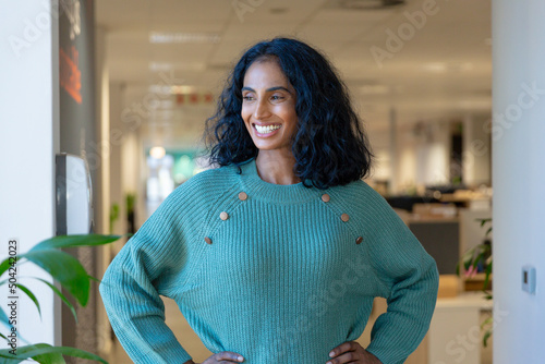 Happy biracial young female advisor standing with hands on hips looking away at modern workplace