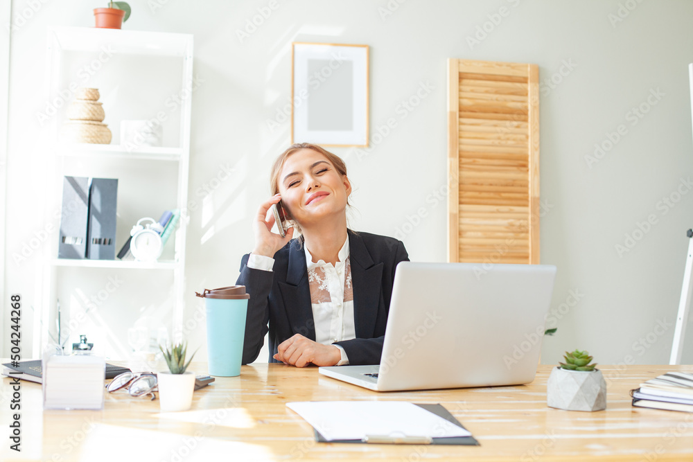 Smiling businesswoman in suit using laptop while talking on mobile phone at desk in home office Smiling businesswoman in suit using laptop while talking on mobile phone at desk in home office