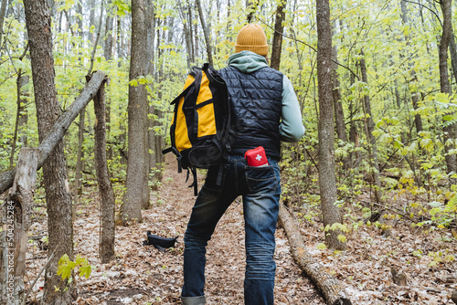 A man stands in the woods from behind, a first aid kit is in his trouser pocket, a tourist with a backpack, a traveler in the woods, a yellow bag hanging on his shoulder, hiking equipment