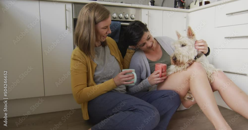 Caucasian lesbian couple holding coffee cups playing with their dog sitting in the kitchen at home Caucasian lesbian couple holding coffee cups playing with their dog sitting in the kitchen at home