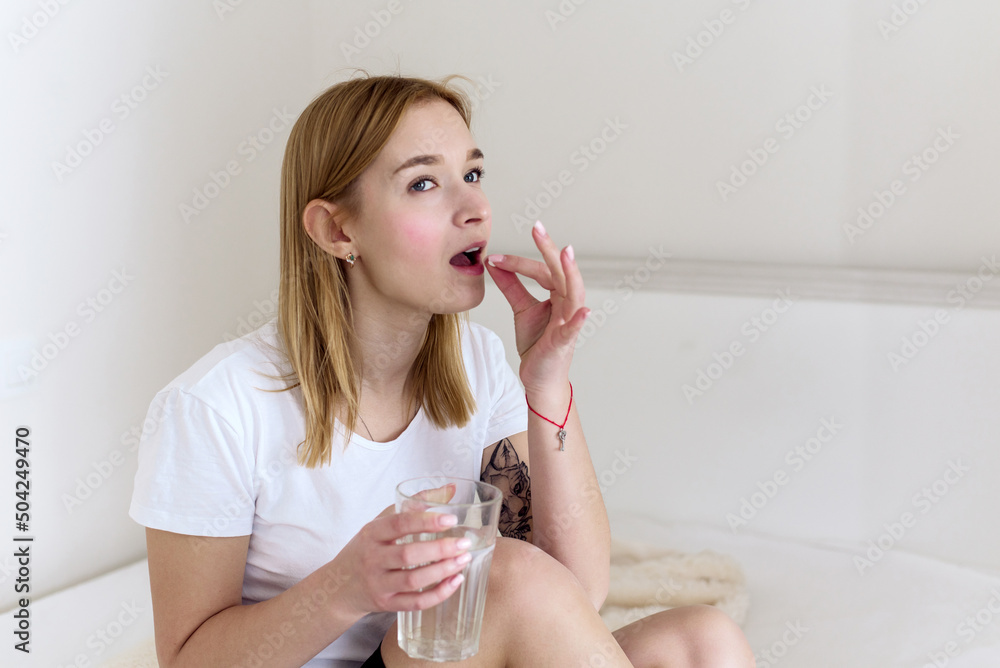Young woman with glass of water taking sleeping pills and not feeling well in bed, copy space. Using medical drug at home. Vitamin or supplement Young woman with glass of water taking sleeping pills and not feeling well in bed, copy space. Using medical drug at home. Vitamin or supplement