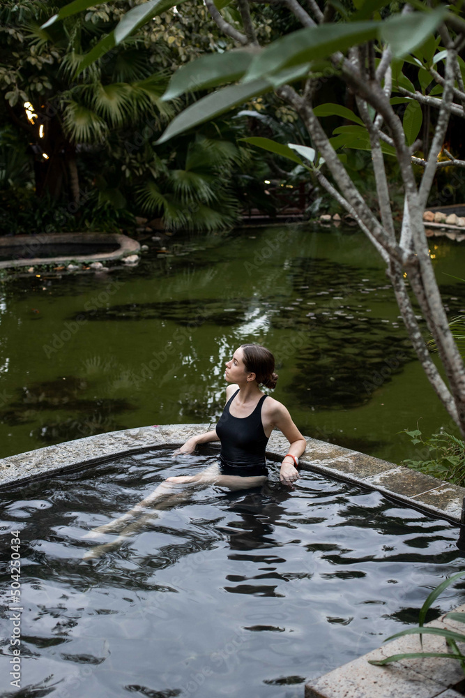 Fototapeta premium A young and beautiful girl in a black swimsuit is relaxing in the thermal springs pool.