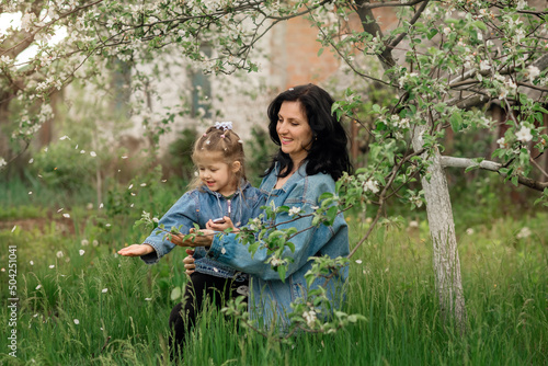 Mother and daughter  are sitting on the grass in a blooming apple orchard and touching falling flowers 