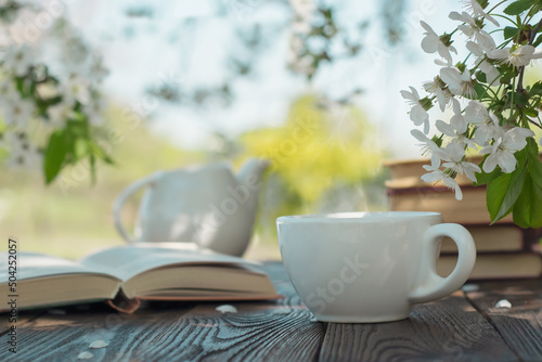 Cup with tea, books, teapot, on a wooden textured table on a background of flowering tree.