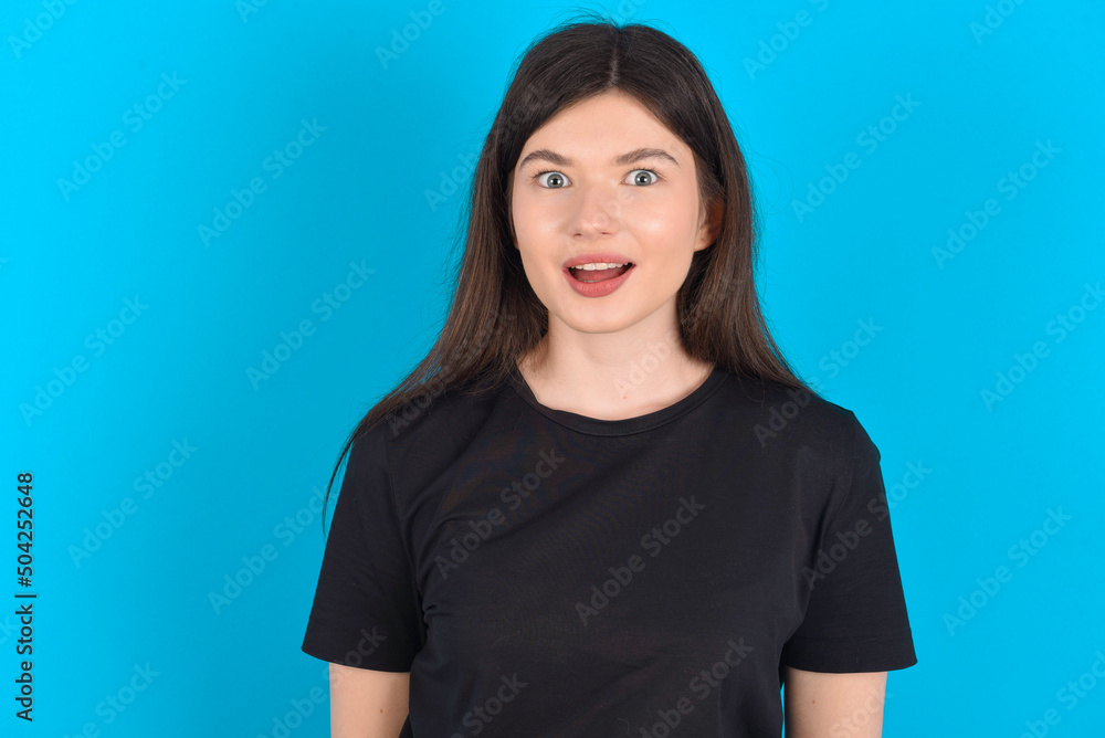 young caucasian woman wearing black T-shirt over blue background having stunned and shocked look, with mouth open and jaw dropped exclaiming: Wow, I can't believe this. Surprise and shock young caucasian woman wearing black T-shirt over blue background having stunned and shocked look, with mouth open and jaw dropped exclaiming: Wow, I can't believe this. Surprise and shock
