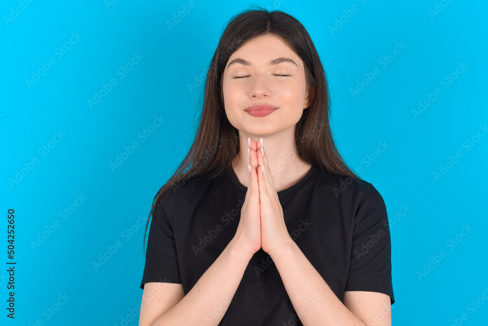 Indoor closeup of young caucasian woman wearing black T-shirt over blue background practicing yoga and meditation, holding palms together in namaste, looking calm, relaxed and peaceful. Indoor closeup of young caucasian woman wearing black T-shirt over blue background practicing yoga and meditation, holding palms together in namaste, looking calm, relaxed and peaceful.