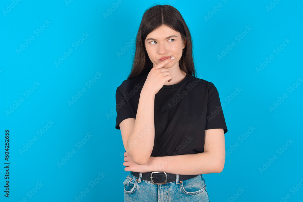 Shot of contemplative thoughtful young caucasian woman wearing black T-shirt over blue background keeps hand under chin, looks thoughtfully upwards. Shot of contemplative thoughtful young caucasian woman wearing black T-shirt over blue background keeps hand under chin, looks thoughtfully upwards.