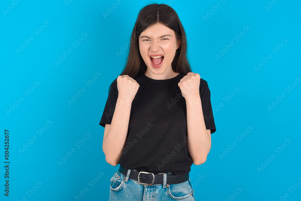young caucasian woman wearing black T-shirt over blue background rejoicing success and victory clenching his fists with joy being happy to achieve her aim and goals. Positive emotions, feelings. young caucasian woman wearing black T-shirt over blue background rejoicing success and victory clenching his fists with joy being happy to achieve her aim and goals. Positive emotions, feelings.