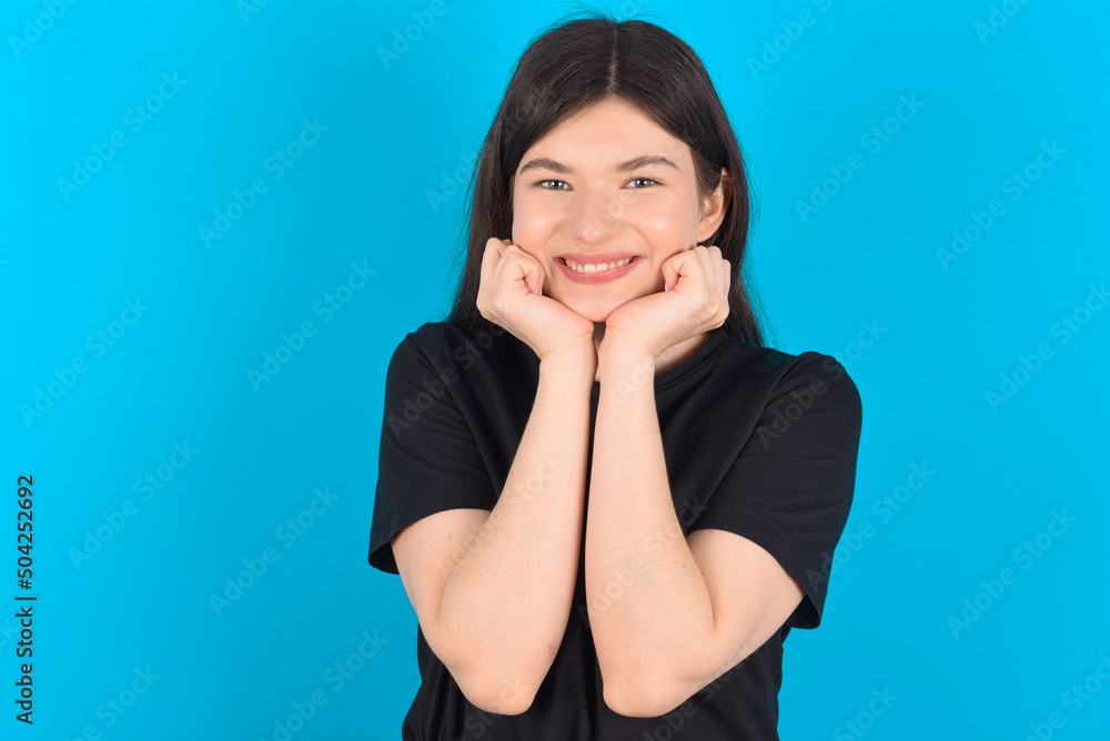 Dreamy young caucasian woman wearing black T-shirt over blue background keeps hands pressed together under chin, looks with happy expression, has toothy smile. Dreamy young caucasian woman wearing black T-shirt over blue background keeps hands pressed together under chin, looks with happy expression, has toothy smile.