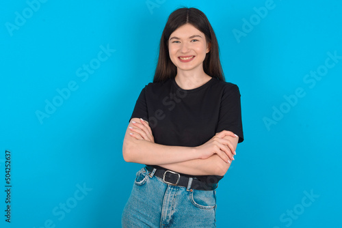young caucasian woman wearing black T-shirt over blue background being happy smiling and crossed arms looking confident at the camera. Positive and confident person. © Jihan