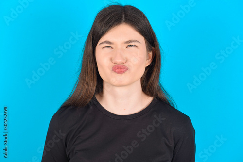young caucasian woman wearing black T-shirt over blue background crosses eyes, puts lips, makes grimace with awkward expression has fun alone, plays fool.
