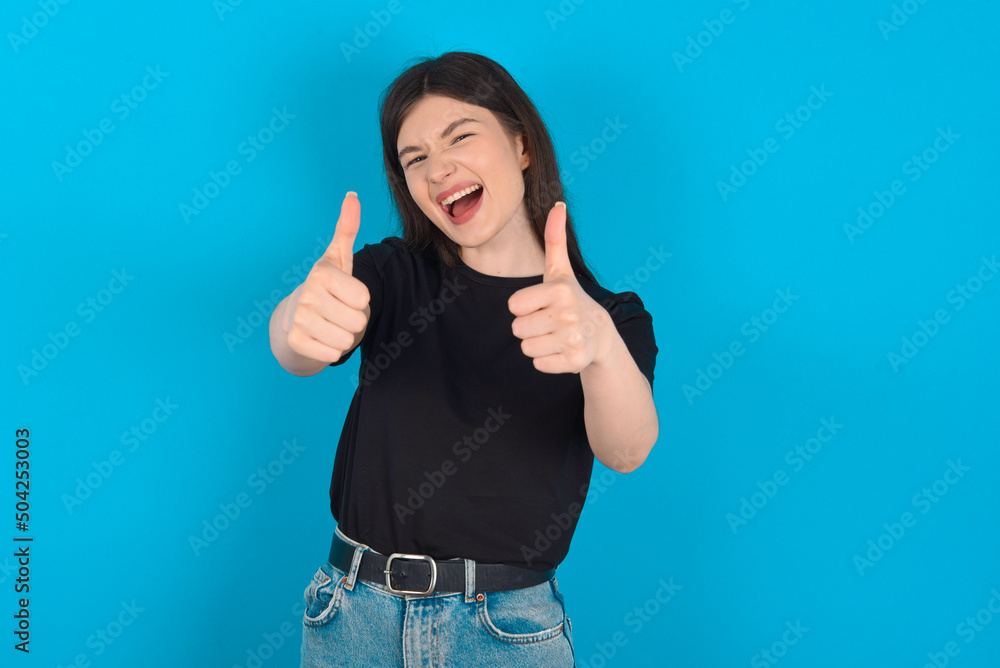 young caucasian woman wearing black T-shirt over blue background making positive gesture with thumbs up smiling and happy for success. Looking at the camera, winner gesture. young caucasian woman wearing black T-shirt over blue background making positive gesture with thumbs up smiling and happy for success. Looking at the camera, winner gesture.