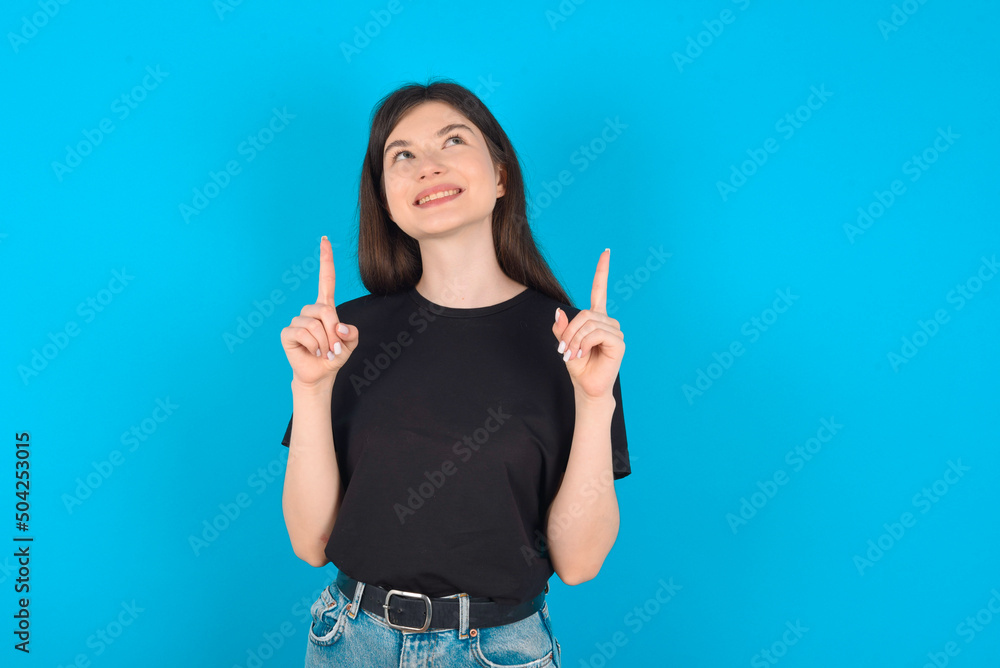 Successful friendly looking young caucasian woman wearing black T-shirt over blue background exclaiming excitedly, pointing both index fingers up, indicating something. Successful friendly looking young caucasian woman wearing black T-shirt over blue background exclaiming excitedly, pointing both index fingers up, indicating something.