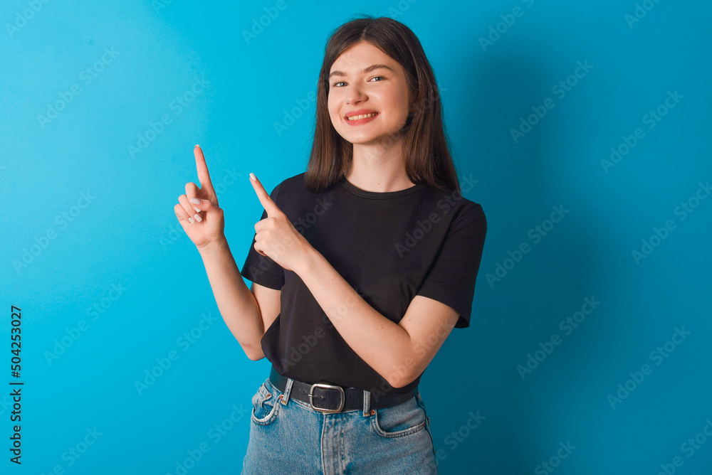 Positive young caucasian woman wearing black T-shirt over blue background with beaming smile pointing with two fingers and looking on empty copy space. Advertisement concept.