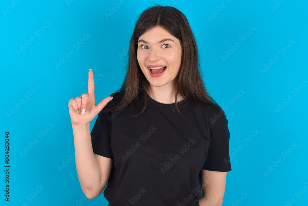 Pleasant looking young caucasian woman wearing black T-shirt over blue background has clever expression, raises one finger, remembers himself not to forget tell important thing. Pleasant looking young caucasian woman wearing black T-shirt over blue background has clever expression, raises one finger, remembers himself not to forget tell important thing.