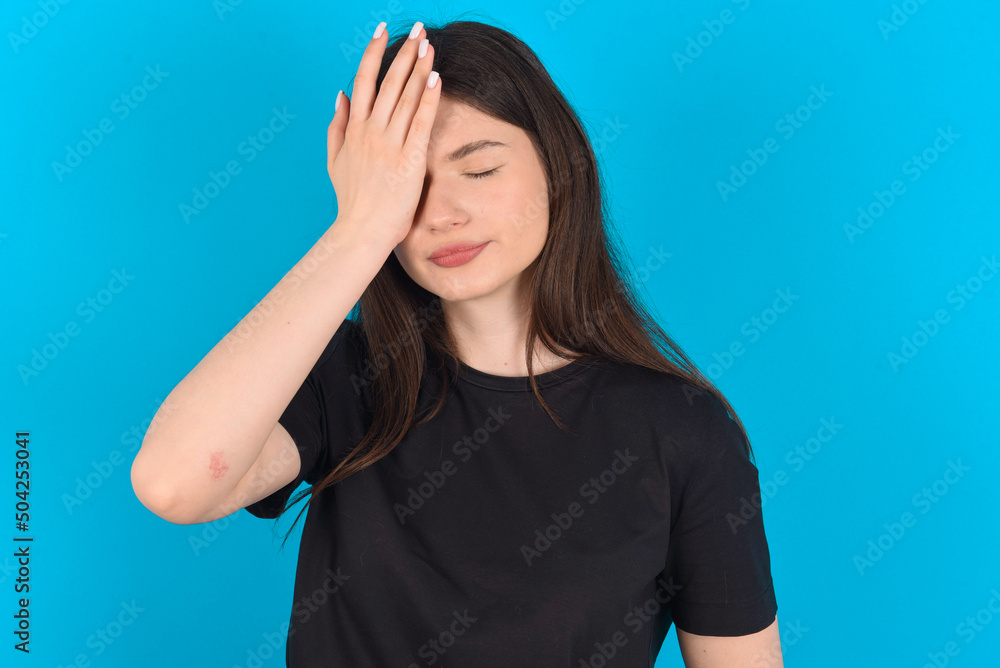 Frustrated young caucasian woman wearing black T-shirt over blue background holding hand on forehead being depressed regretting what he did having headache, looking stressful. Frustrated young caucasian woman wearing black T-shirt over blue background holding hand on forehead being depressed regretting what he did having headache, looking stressful.