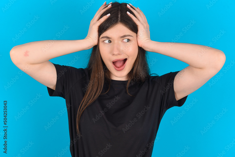 Horrible, stress, shock. Portrait emotional crazy young caucasian woman wearing black T-shirt over blue background clasping head in hands. Emotions, facial expression concept.
