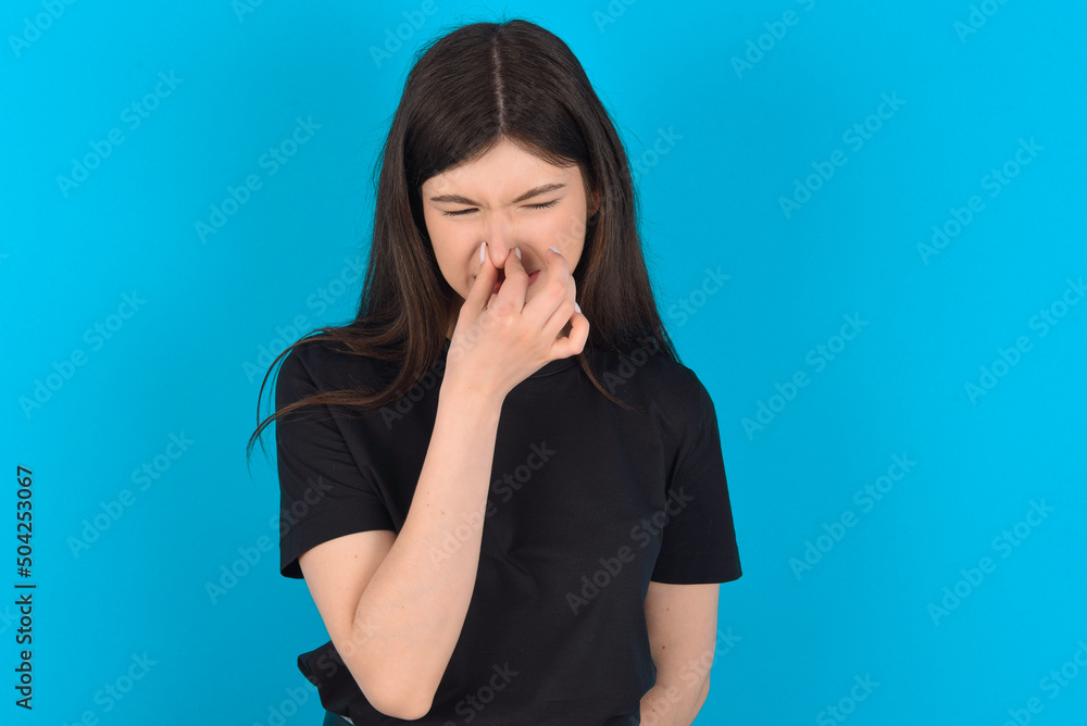 young caucasian woman wearing black T-shirt over blue background , holding his nose because of a bad smell. young caucasian woman wearing black T-shirt over blue background , holding his nose because of a bad smell.