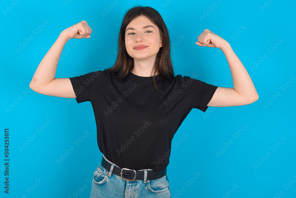 Waist up shot of young caucasian woman wearing black T-shirt over blue raises arms to show muscles feels confident in victory, looks strong and independent, smiles positively at camera. Sport concept. Waist up shot of young caucasian woman wearing black T-shirt over blue raises arms to show muscles feels confident in victory, looks strong and independent, smiles positively at camera. Sport concept.