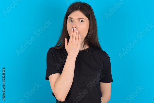 Oh! I think I said it! Close up portrait young caucasian woman wearing black T-shirt over blue background cover open mouth by hand palm, look at camera with big eyes.