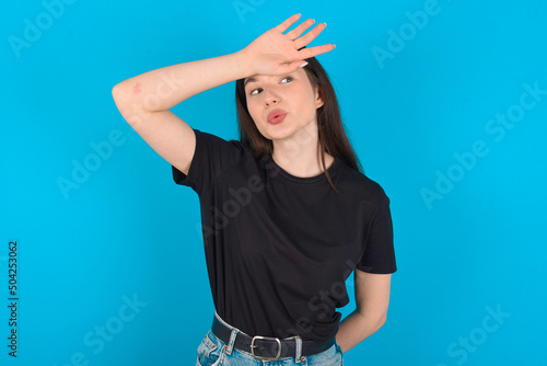 young caucasian woman wearing black T-shirt over blue background wiping forehead with hand making phew gesture, expressing relief feels happy that he prevented huge disaster. It was close enough photo