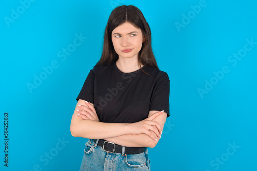 Displeased young caucasian woman wearing black T-shirt over blue background with bad attitude, arms crossed looking sideways. Negative human emotion facial expression feelings. © Jihan
