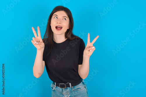 Isolated shot of cheerful young caucasian woman wearing black T-shirt over blue background makes peace or victory sign with both hands  feels cool.