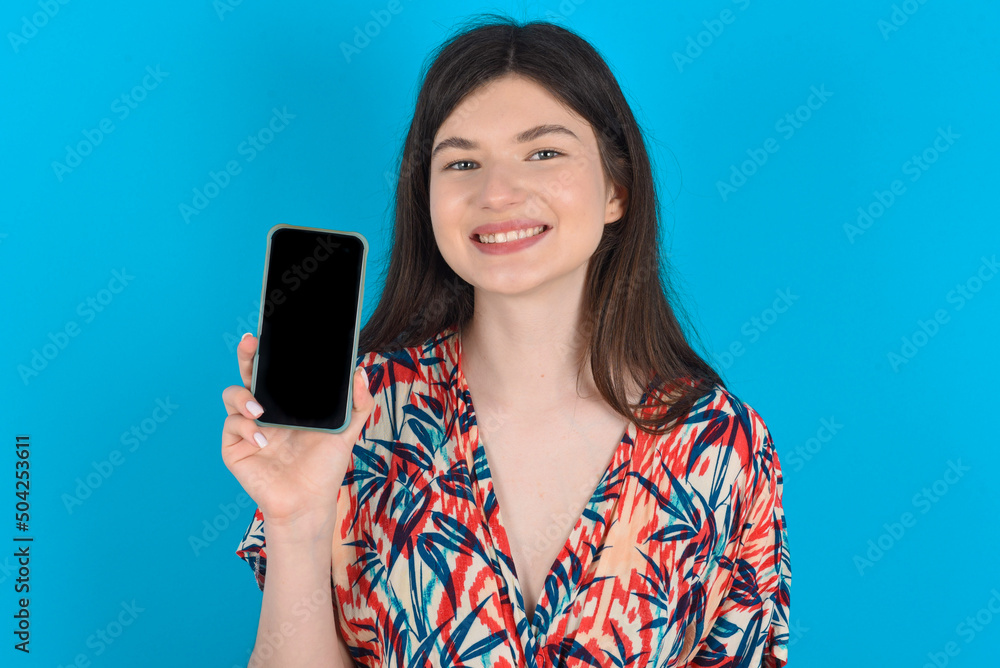Smiling young caucasian woman wearing floral dress over blue background showing  empty phone screen. Advertisement and communication concept.
