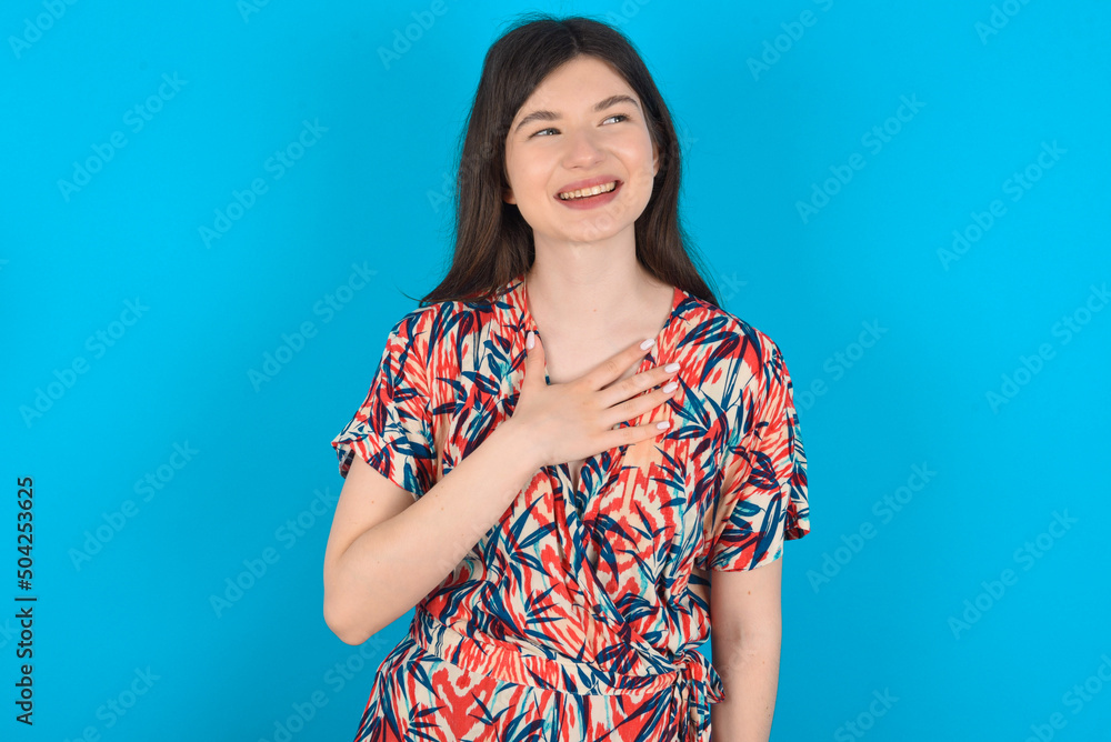 Joyful young caucasian woman wearing floral dress over blue background expresses positive emotions recalls something funny keeps hand on chest and giggles happily. Joyful young caucasian woman wearing floral dress over blue background expresses positive emotions recalls something funny keeps hand on chest and giggles happily.
