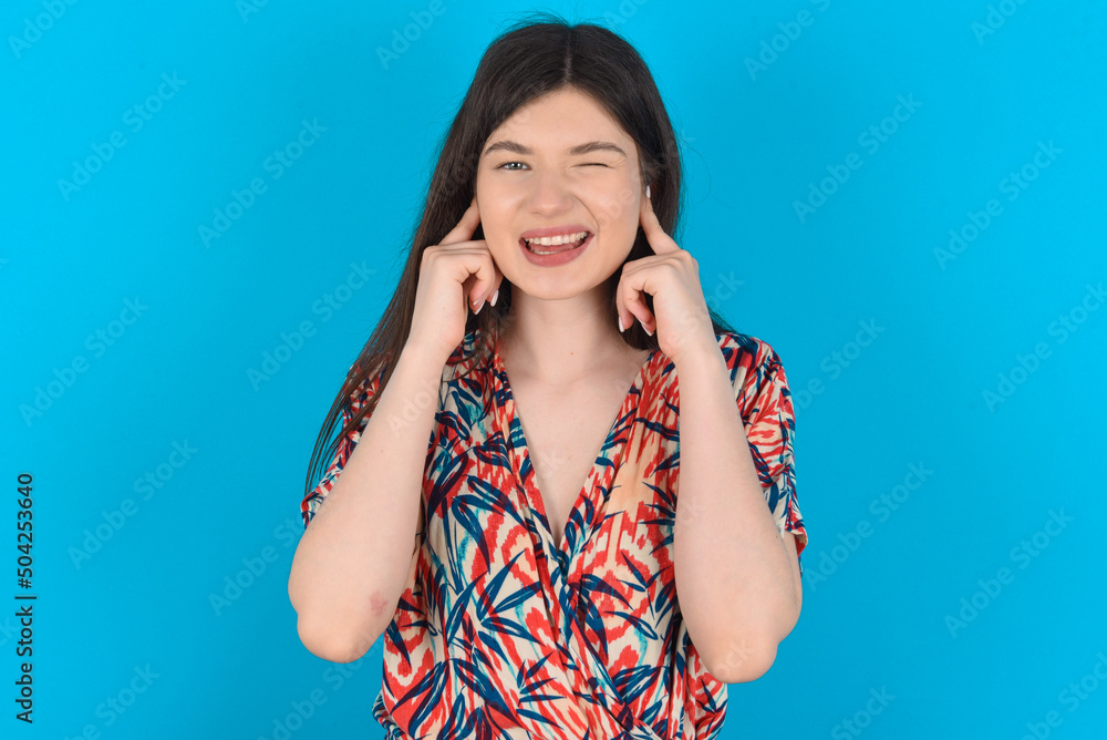 Happy young caucasian woman wearing floral dress over blue background ignores loud music and plugs ears with fingers asks to turn off sound Happy young caucasian woman wearing floral dress over blue background ignores loud music and plugs ears with fingers asks to turn off sound