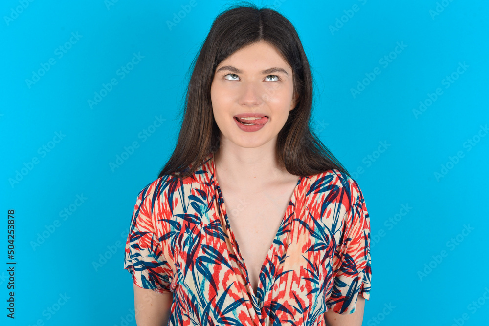 Fototapeta premium Funny young caucasian woman wearing floral dress over blue background makes grimace and crosses eyes plays fool has fun alone sticks out tongue.