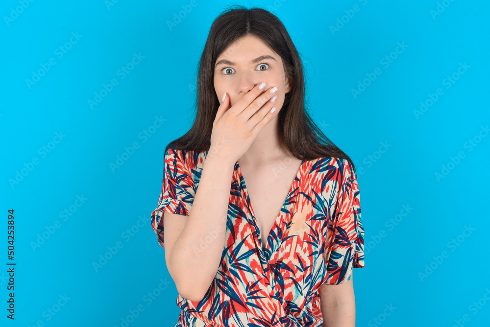 young caucasian woman wearing floral dress over blue background covers mouth and looks with wonder at camera, cannot believe unexpected rumors. young caucasian woman wearing floral dress over blue background covers mouth and looks with wonder at camera, cannot believe unexpected rumors.