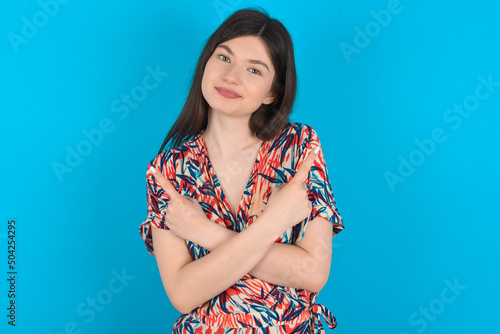 young caucasian woman wearing floral dress over blue background crosses arms and points at different sides hesitates between two items or variants. Needs help with decision