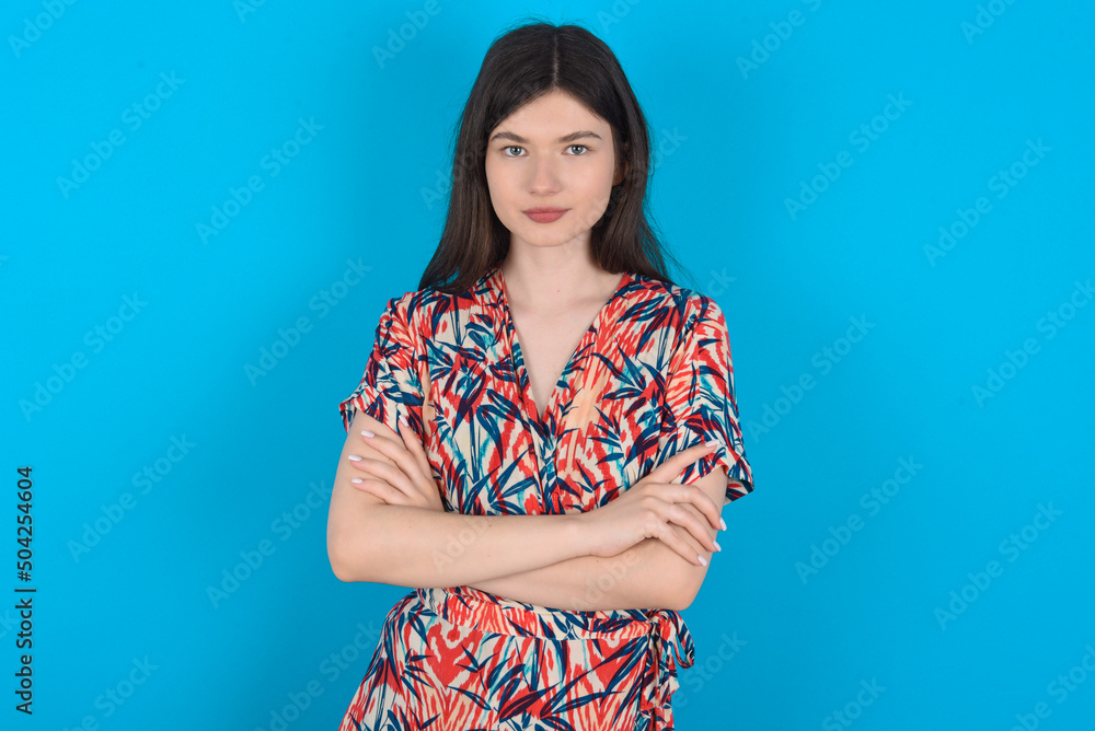 Confident young caucasian woman wearing floral dress over blue background with arms crossed looking to the camera Confident young caucasian woman wearing floral dress over blue background with arms crossed looking to the camera