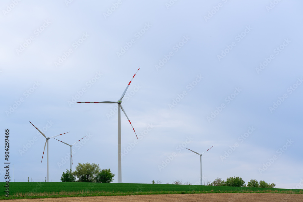 Fototapeta premium Wind turbines on a blue background stand on a green field with rare green trees on a spring day, green energy saves nature and beauty