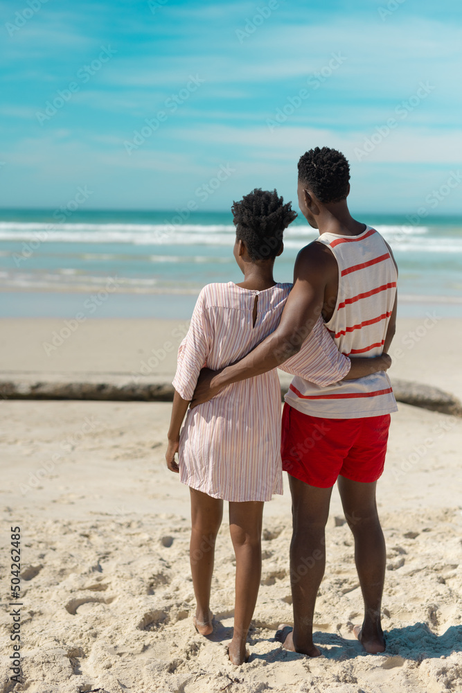 Rear view of african american young couple with arms around looking at sea while standing on beach Rear view of african american young couple with arms around looking at sea while standing on beach
