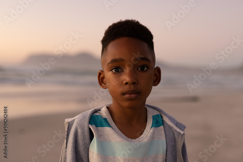 Portrait of african american cute boy wearing jacket standing at beach against clear sky at sunset