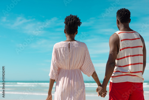 Rear view of african american young couple holding hands and looking at sea while standing on beach
