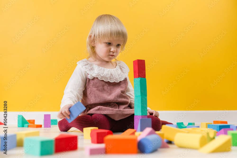 A little serious girl in a dress is sitting on the floor and building a tower of multicolored designer blocks A little serious girl in a dress is sitting on the floor and building a tower of multicolored designer blocks