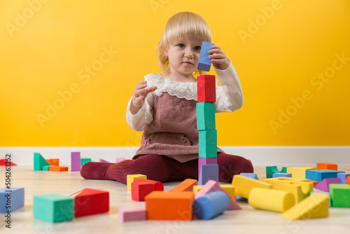 A little focused girl sits on the floor and puts a blue cube on a tower of colored blocks of the constructor